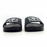 Image of black slide sandals against a white background. The middle of the strap on the slides feature the tinashe 333 logo in white. The logo is a 3 facing the right way, mirrored by a 3 facing the opposite way, connected to a large 3 that is sideways and across the bottom of the other two 3's, forming a square shaped logo. 