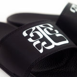 up close Image of black slide sandals against a white background. The middle of the strap on the slides feature the tinashe 333 logo in white. The logo is a 3 facing the right way, mirrored by a 3 facing the opposite way, connected to a large 3 that is sideways and across the bottom of the other two 3's, forming a square shaped logo.