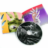 Image of a cd booklet and cd against a white background. The cd and booklet are for tinashe's album "333". the cd is grey and white with abstract imagery. the booklet shows colorful pages- the left is blue, green, white and yellow and features an up close graphic of a hand with an eye in the center. The right page is a pink, grey and purple gradient and features a white hand with an eye in the center of it and another hand wrapped around the wrist of the eyeball hand.